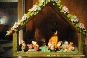 Căsuță decorativă din marțipan, cu flori pe acoperiș și figurine mici.