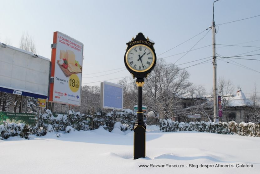 Ceas ornamental negru lângă panouri publicitare, înconjurat de zăpadă.
