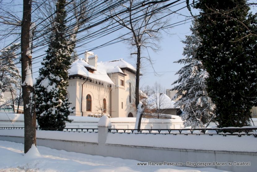Casă deschisă la culoare, cu acoperiș alb, în spatele unui gard și arbori.