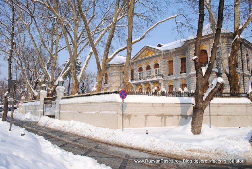 Clădire cu coloane și balcoane, dincolo de un zid alb și trotuar înzăpezit.
