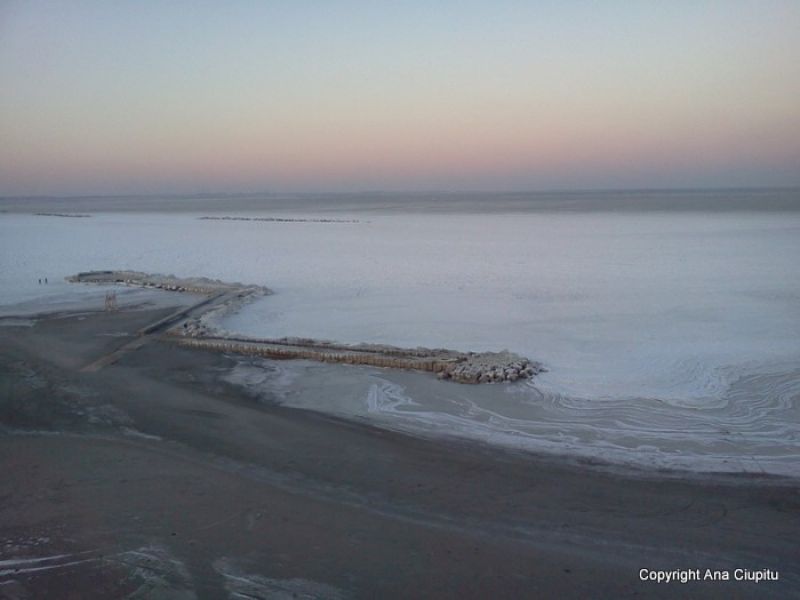 Plajă curbată și mare înghețată, sub o fâșie rozalie de cer.