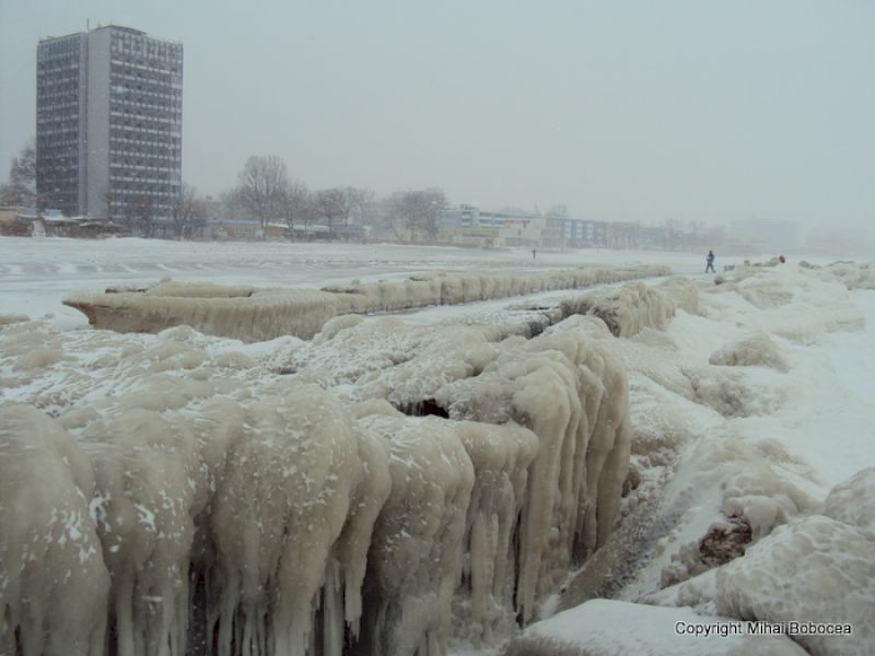 Dig încărcat cu gheață și țurțuri, cu un bloc înalt în fundal.