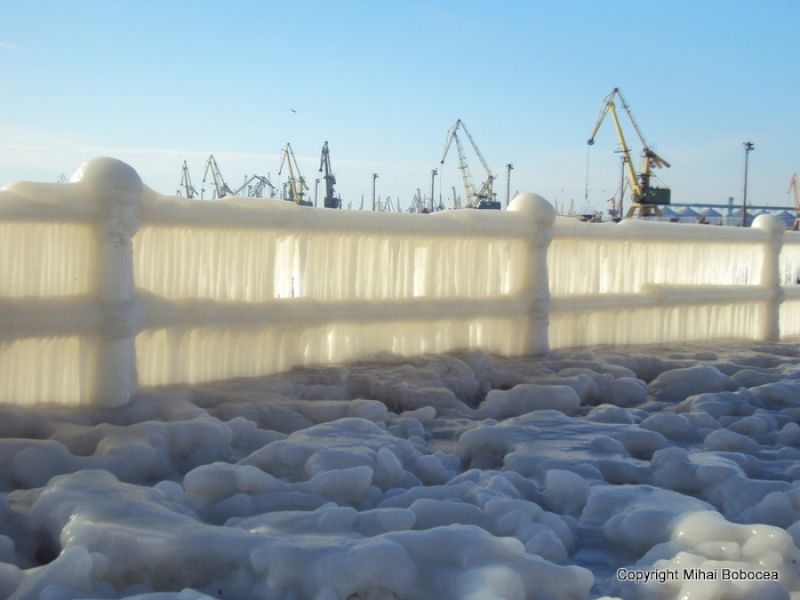 Balustradă acoperită cu țurțuri, în fața macaralelor portuare sub cer albastru.