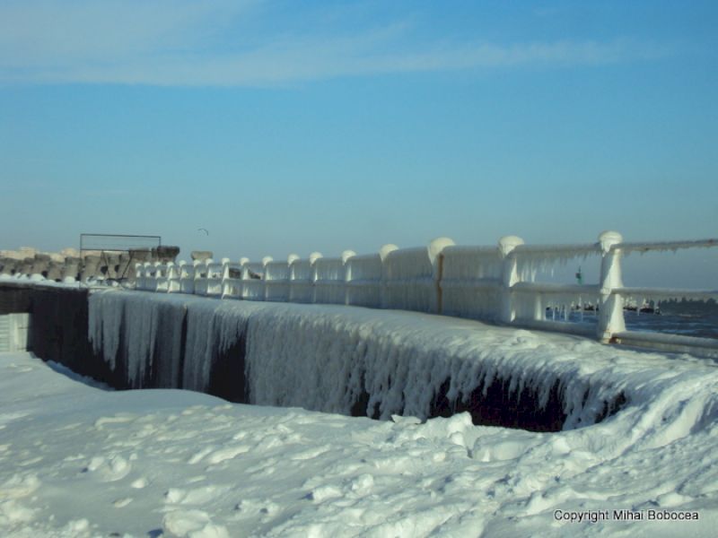 Balustradă albă deasupra unui zid cu țurțuri lungi, sub cer albastru.