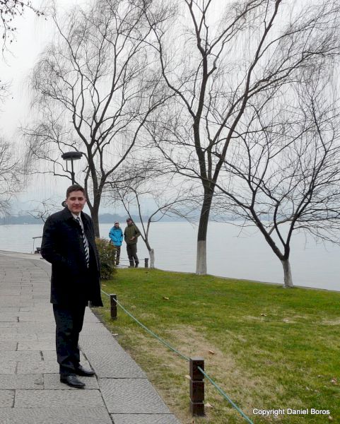 Bărbat în haină neagră pe o alee lângă lac și arbori desfrunziți.