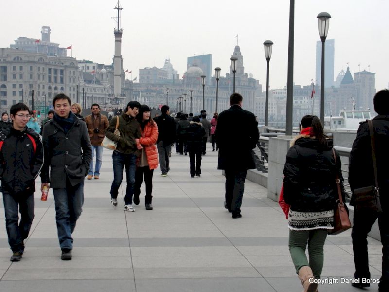 Pietoni pe o promenadă largă, cu felinare și clădiri înalte în fundal.