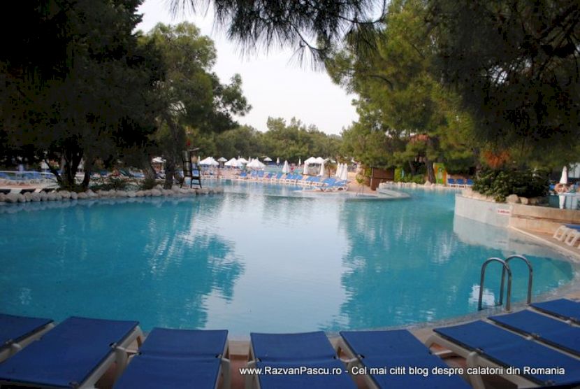 Piscină largă văzută din dreptul șezlongurilor albastre și al scării metalice.