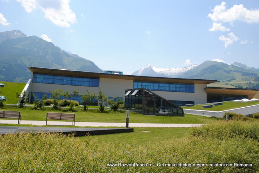 Complex modern cu fațadă vitrată, înconjurat de pajiști și munți.