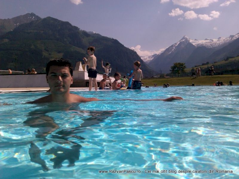 Bărbat înoată într-o piscină exterioară, cu turiști și munți în fundal.