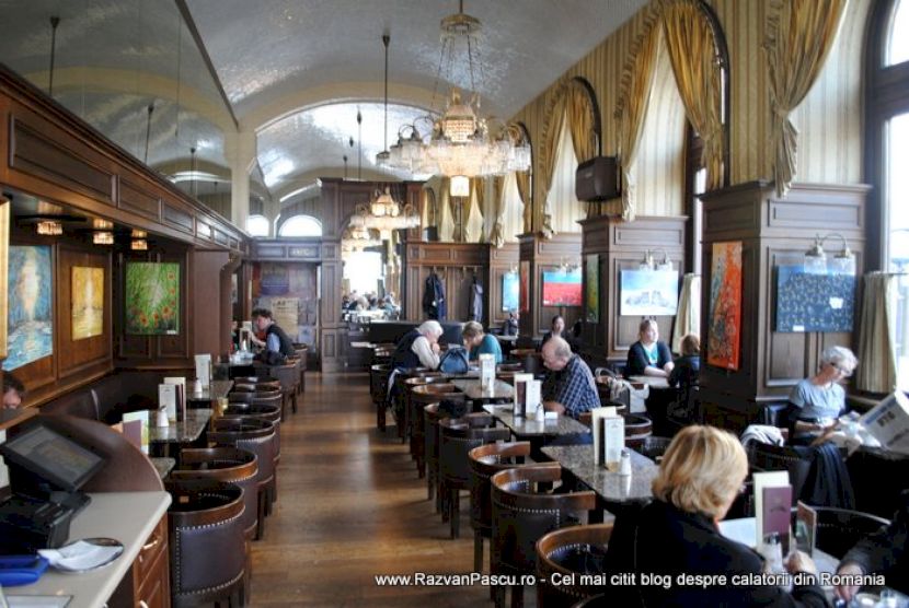 Interior de cafenea cu bolți albe, candelabre și mese ocupate.
