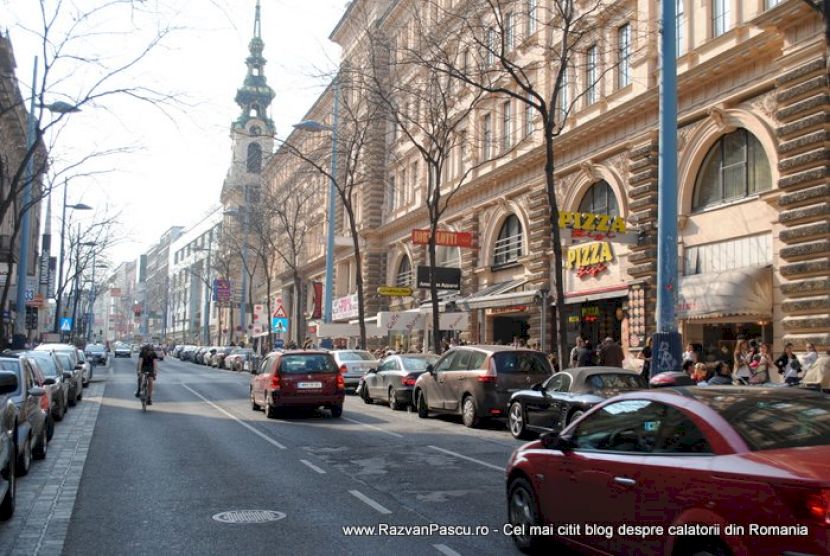 Stradă cu mașini parcate, clădiri istorice și turn în depărtare.