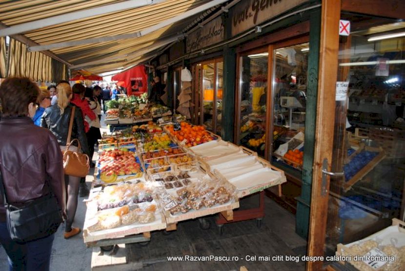 Cumpărători lângă tarabe alimentare acoperite de o copertină dungată.