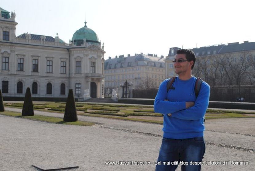 Bărbat în bluză albastră, cu brațele încrucișate în fața unui palat.