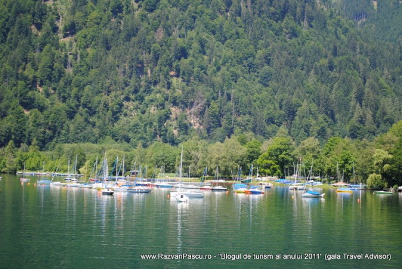 Bărci aliniate pe malul unui lac, sub un versant împădurit.