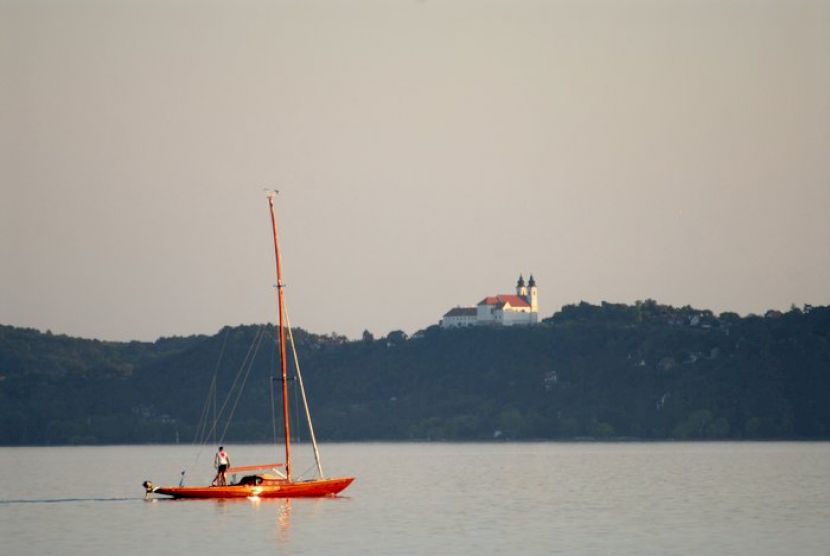 Velier cu cocă roșie pe un lac, în fața dealurilor.