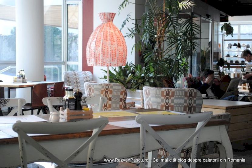 Mese de restaurant lângă ferestre, cu lampă roz și plante înalte.