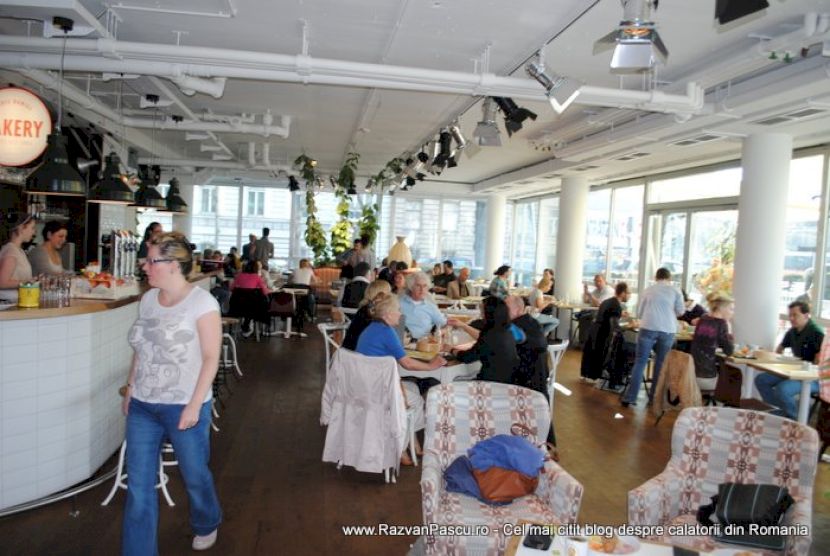 Clienți la mesele restaurantului, lângă tejgheaua albă și ferestrele largi.