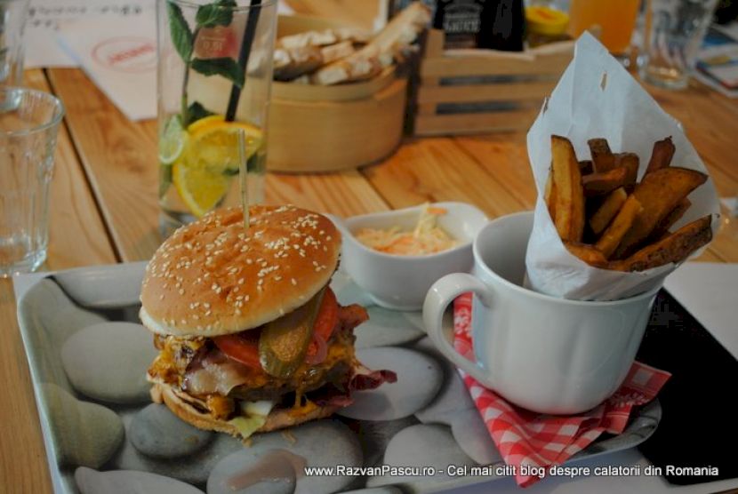 Burger cu chiflă presărată cu susan și cartofi într-o cană albă.