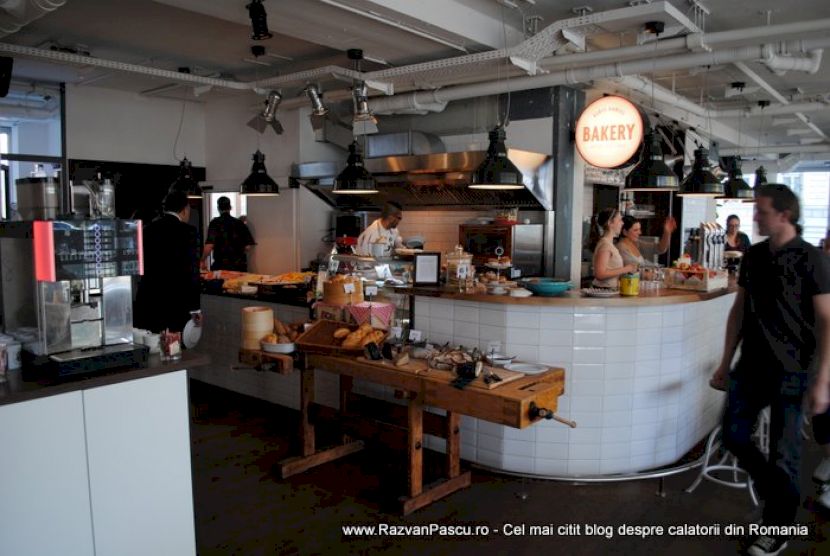Tejghea albă curbată într-o cafenea, cu produse expuse și corpuri industriale.