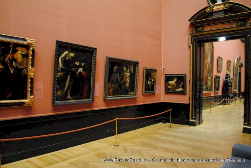 Galerie cu pereți roz, picturi și portal înalt spre sala următoare.