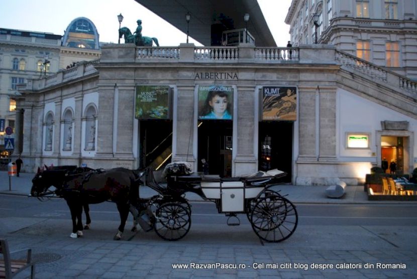 Trăsură trasă de cai, în fața unei clădiri istorice cu statui.