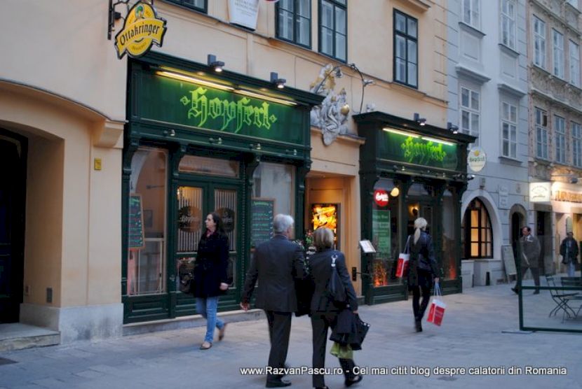 Trecători în fața unui restaurant cu firme verzi și fațadă crem.