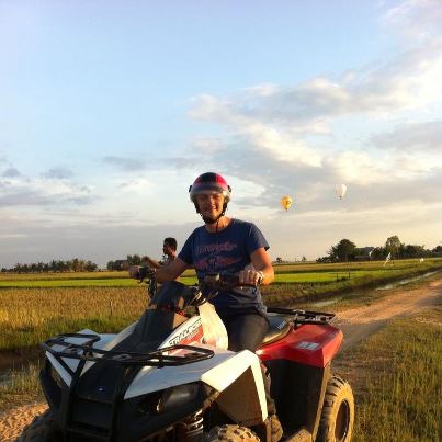 Bărbat cu cască așezat pe un ATV lângă câmpuri.