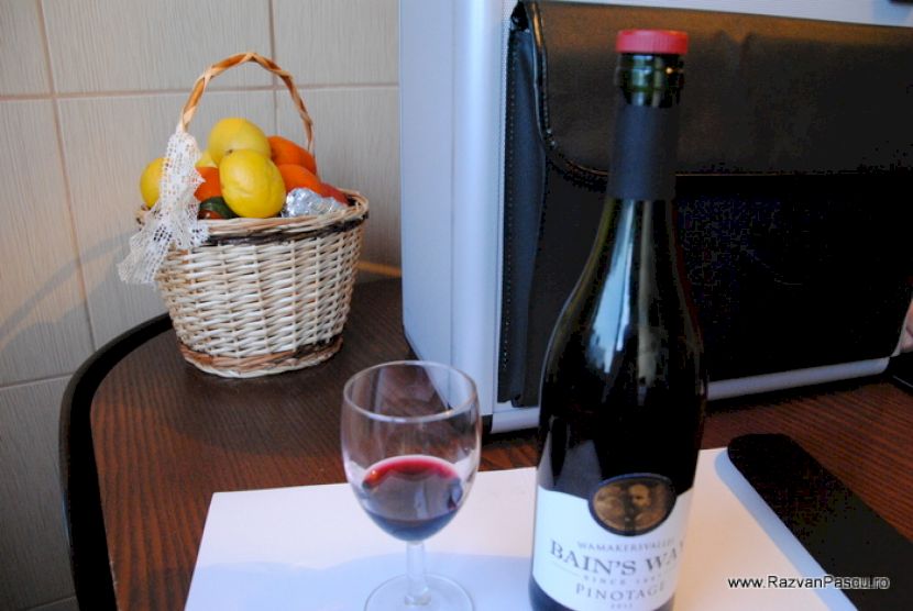Sticlă de vin roșu și pahar lângă un coș cu fructe.