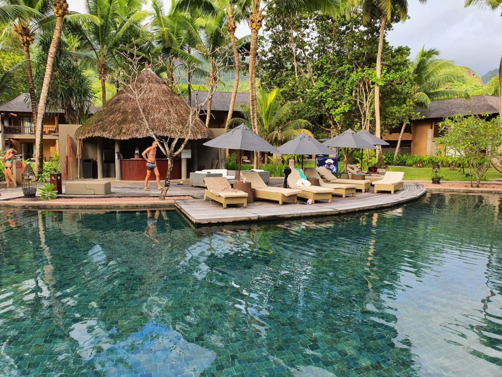 Piscină mărginită de palmieri, șezlonguri și pavilioane cu acoperișuri de paie.