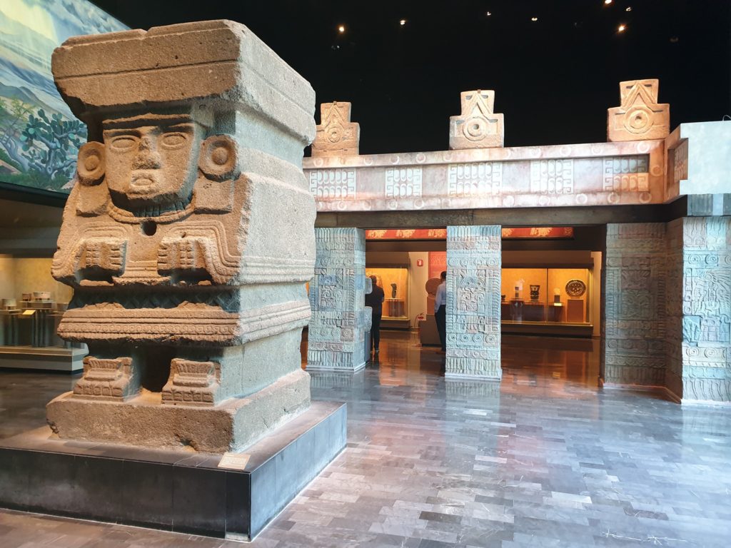 Sculptură din piatră cu chip stilizat, în fața unei structuri muzeale.