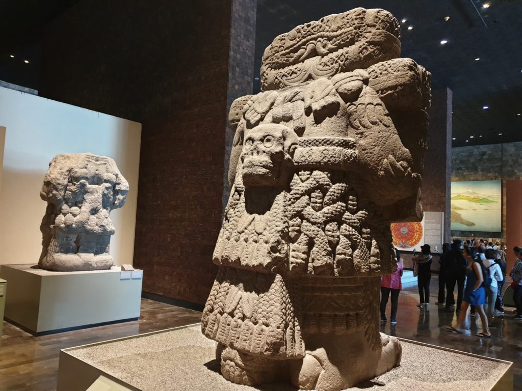 Statuie monumentală din piatră, bogat sculptată, într-o sală de muzeu.