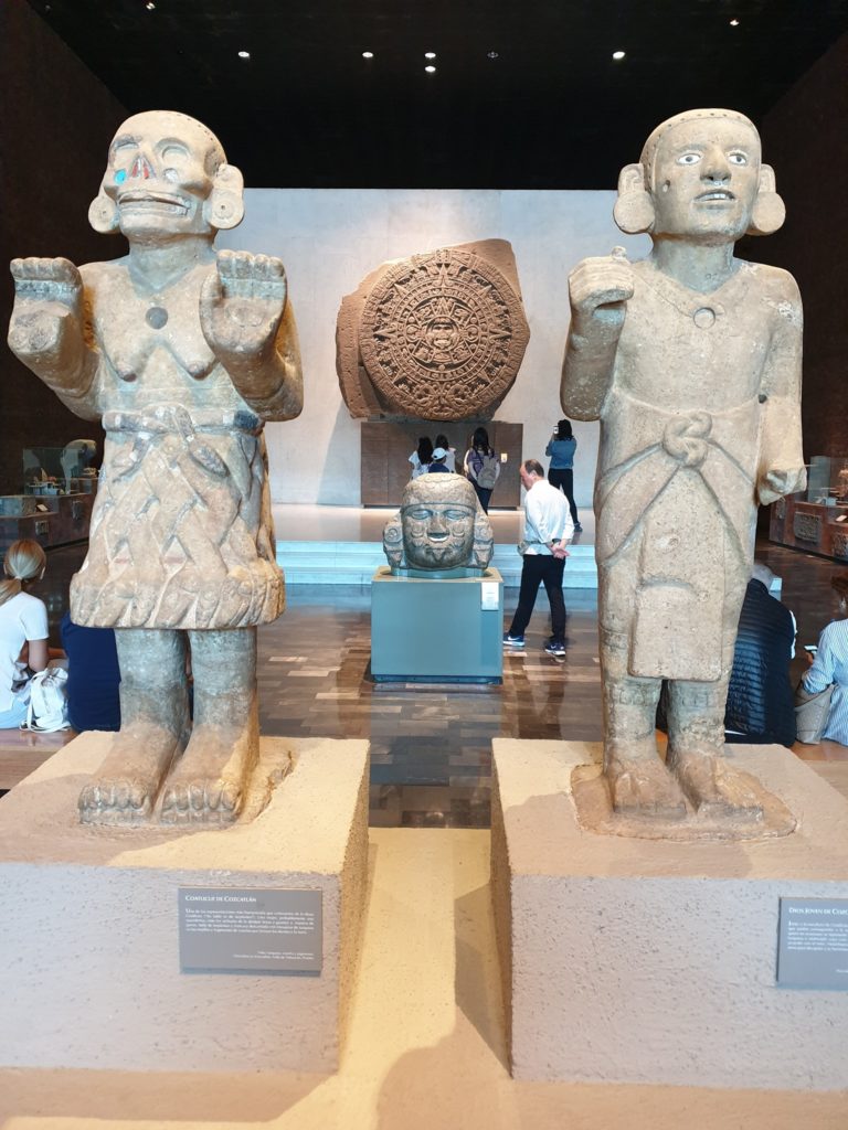 Două statui din piatră expuse în muzeu, lângă un disc sculptat.
