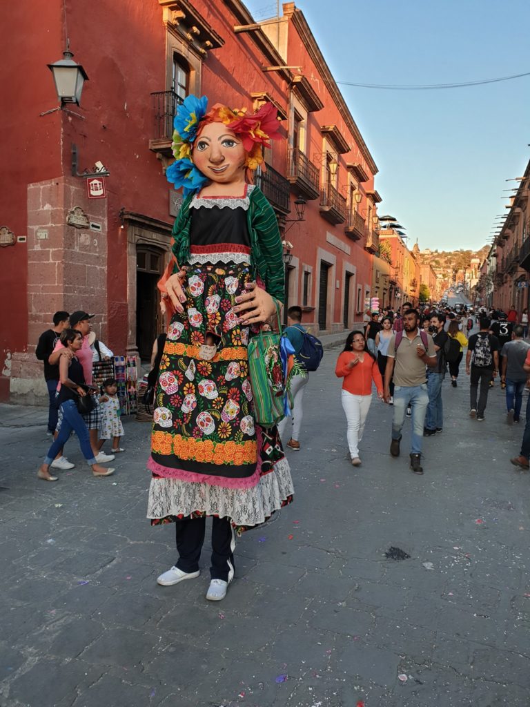 Păpușă uriașă în costum colorat, purtată pe o stradă aglomerată.