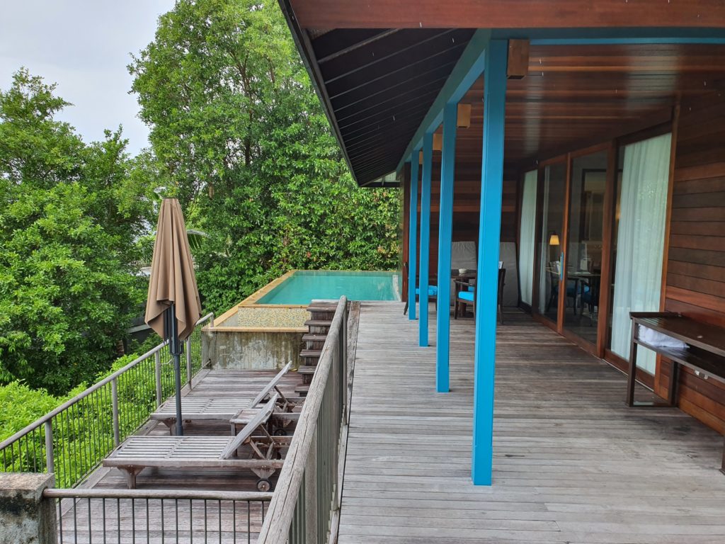 Terasă din lemn cu stâlpi albaștri și piscină între copaci.