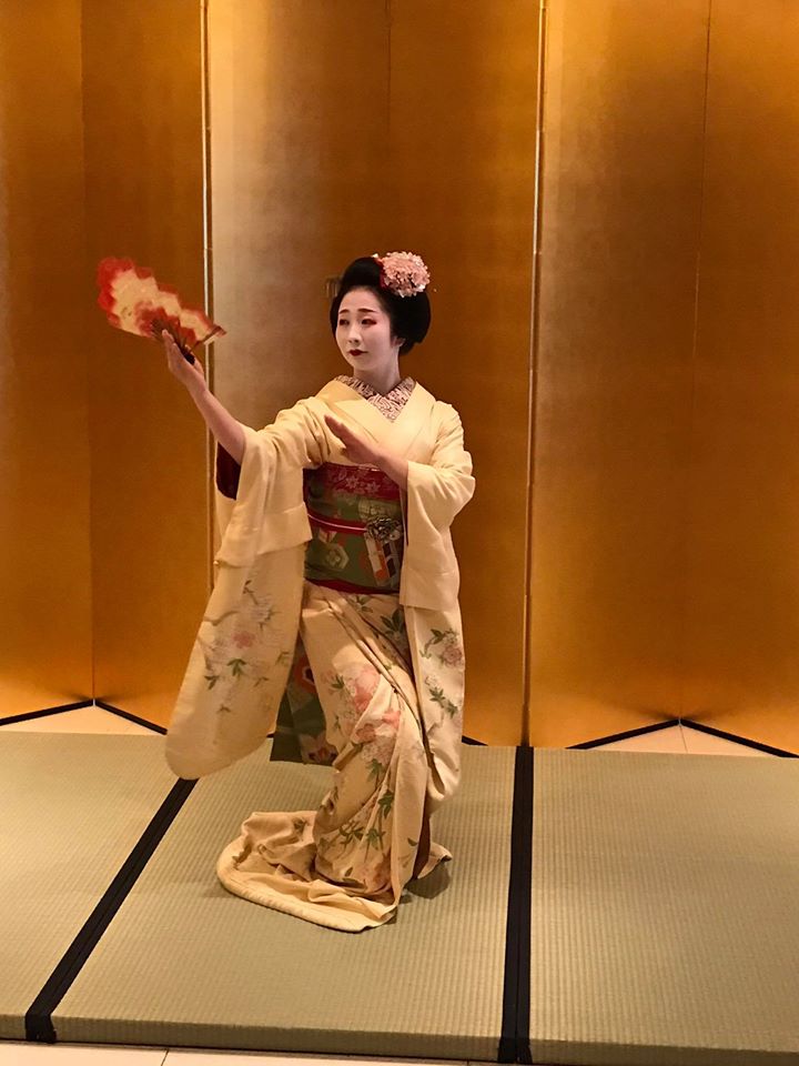 Femeie în kimono dansând cu un evantai în fața unui paravan auriu.