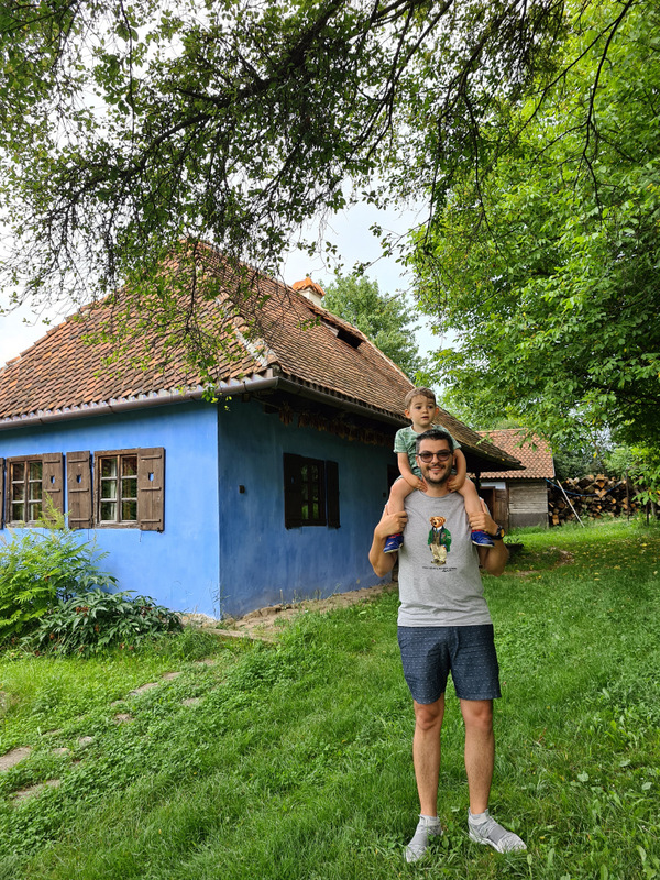 Bărbat cu un copil pe umeri în curtea unei case albastre.