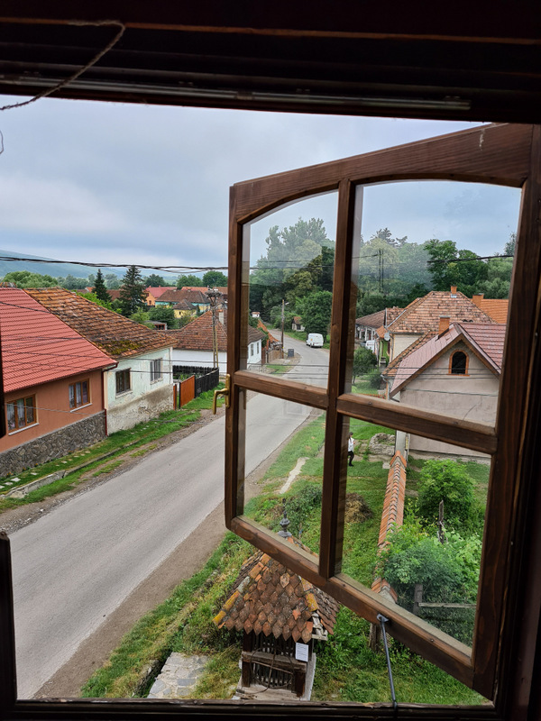 Fereastră deschisă spre ulița satului și acoperișuri de țiglă.
