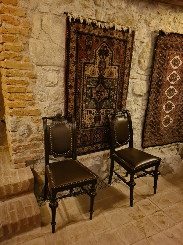 Două scaune din lemn așezate sub covoare decorative pe peretele din piatră.