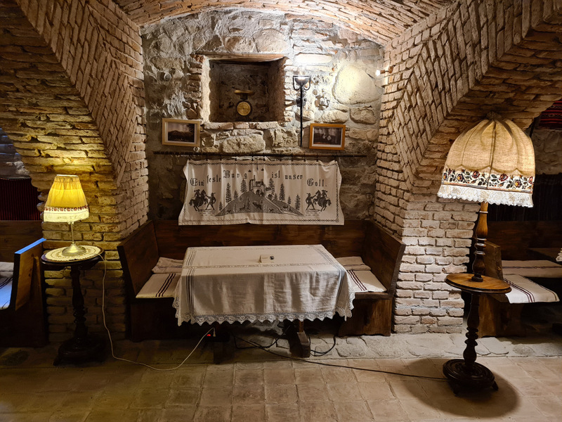 Masă acoperită cu dantelă sub rafturi cu obiecte decorative și tablouri.