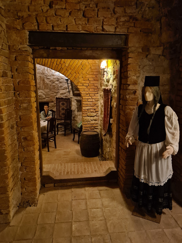 Costum tradițional expus lângă trecerea boltită dintre două încăperi din piatră.