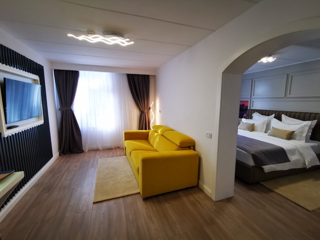 Cameră cu o canapea galbenă și dormitor vizibil printr-o arcadă.