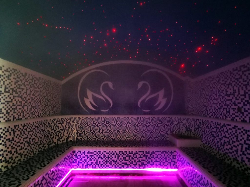 Încăpere cu bănci mozaicate și iluminare violet sub un tavan înstelat.
