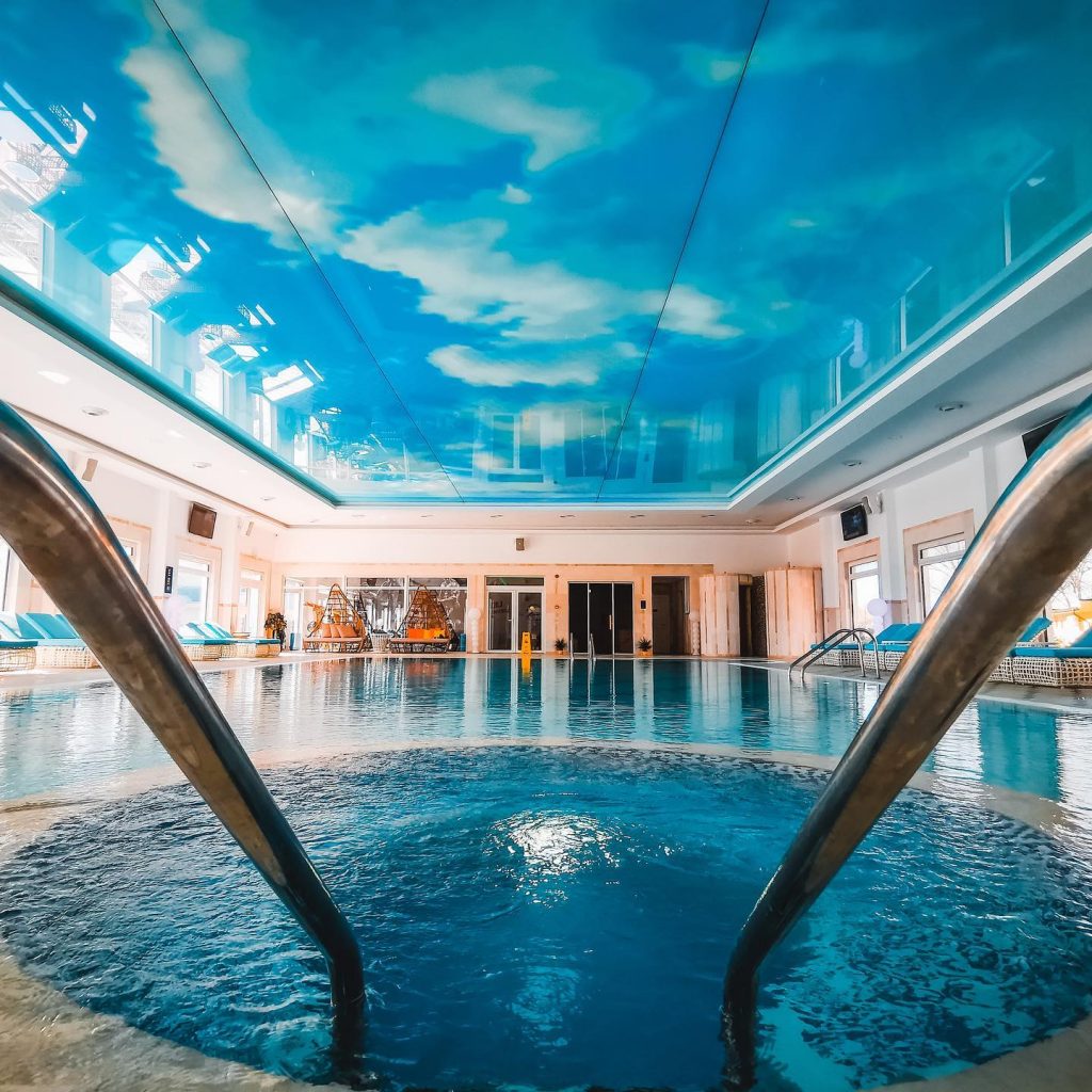 Piscină interioară cu balustrade metalice și tavan pictat cu nori.