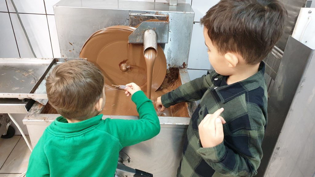 Doi copii privesc ciocolata lichidă dintr-un utilaj.