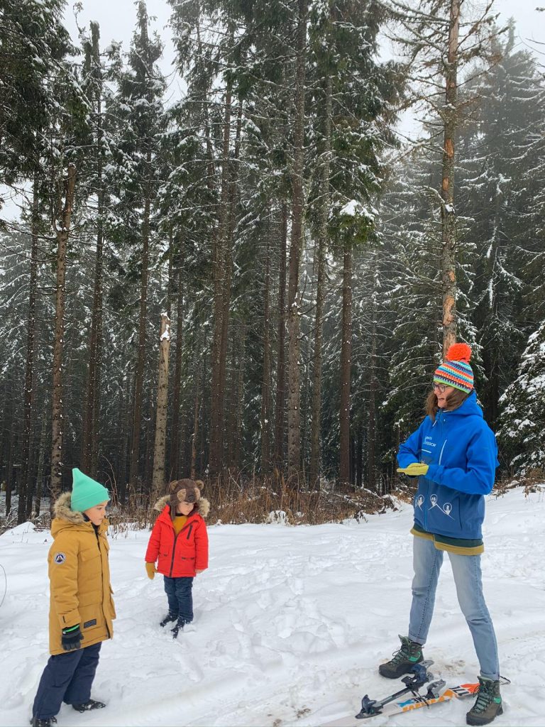 Un adult și doi copii stau pe zăpadă lângă pădure.