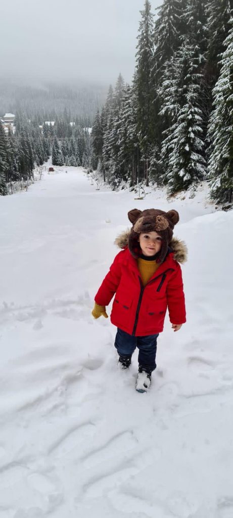 Copil cu geacă roșie pe un drum înzăpezit prin pădure.