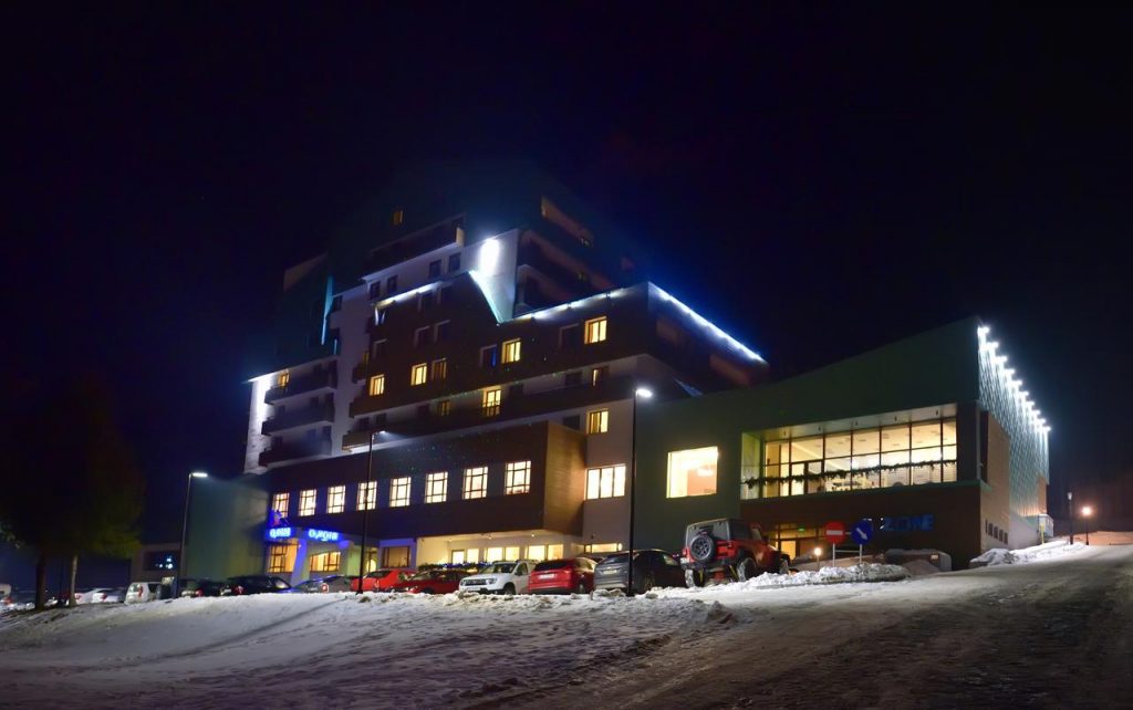 Clădire cu ferestre luminate și contur luminos, fotografiată noaptea pe zăpadă.