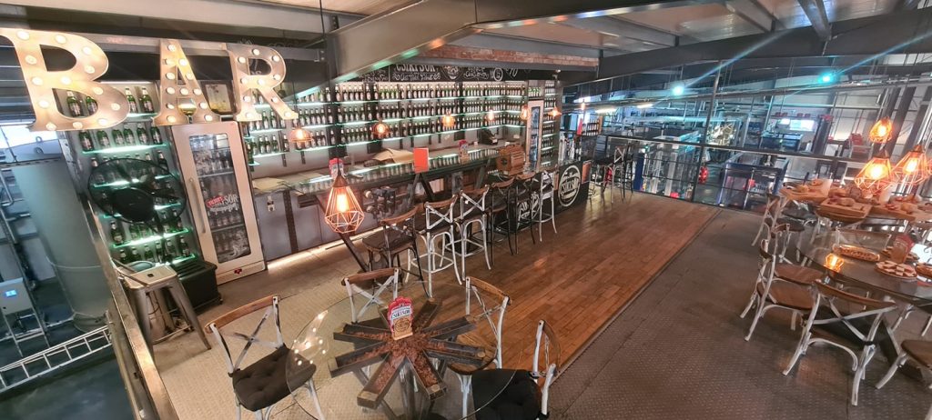 Interior de bar cu tejghea, scaune înalte și instalații metalice.