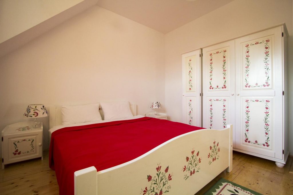 Dormitor cu pat alb pictat, cuvertură roșie și dulap decorat.