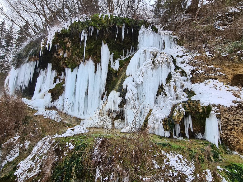 Perete stâncos acoperit cu țurțuri, sub arbori desfrunziți.